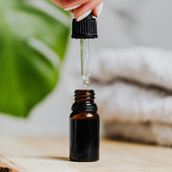 Hand using a dropper to add  diffuser oil to an amber bottle on a wooden surface with a blurred natural background